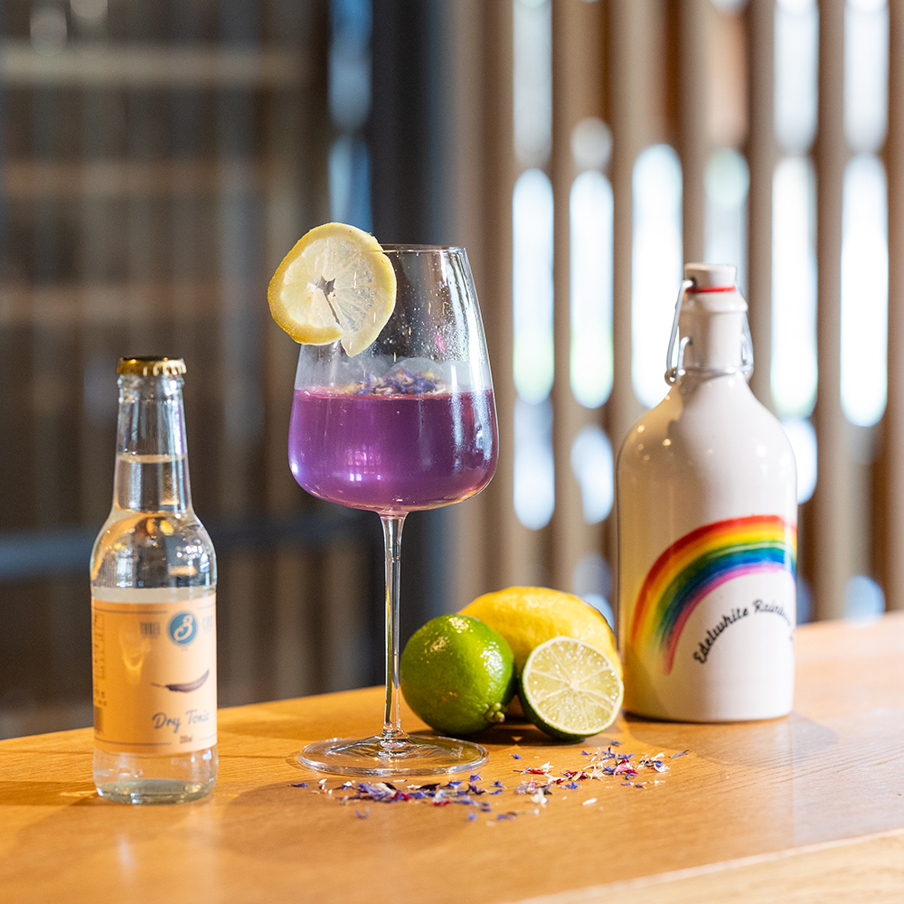 Auf einem Tisch befinden sich ein Weinglas, eine Flasche Limonade und Edelwhite Rainbow Gin & Tonic.